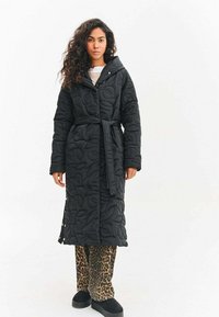 Žena stojaca na bielom pozadí, oblečená v dlhej čiernej prešívanej bunde s opaskom, nohaviciach s leopardím vzorom a čiernych topánkach.