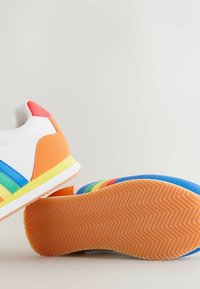 Valkoiset tennarit, joissa oranssi kanta, sivuilla sateenkaarenväriset raidat ja oranssit kuvioidut kumipohjat, esillä vaaleaa taustaa vasten.