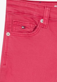 Jeans de mezclilla rojos con un pequeño bolsillo frontal, cierre con botón plateado y costuras en contraste. La tela presenta un acabado suave y texturizado.