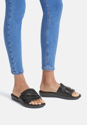 Black slide sandals with textured, layered upper. Low, cushioned sole and open toe design paired with blue skinny jeans.