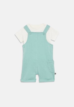 Svetlomodré bavlnené overalové kraťasy s textúrovanou látkou, nastaviteľnými ramienkami, dvoma prednými gombíkmi a bočným vreckom. Pod tým biela košeľa s krátkym rukávom.