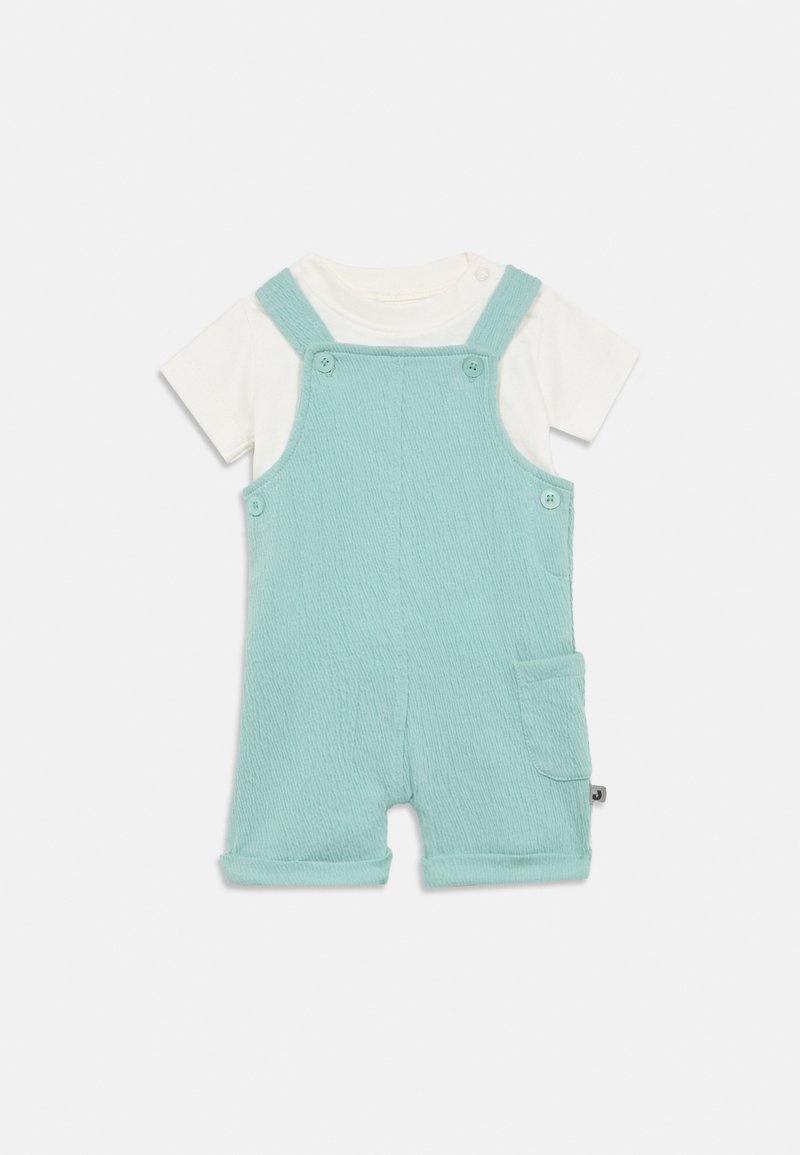 Ανοιχτό μπλε βαμβακερό ολόσωμο σορτς με υφή ύφασμα, ρυθμιζόμενους ιμάντες, δύο κουμπιά μπροστά και μια πλαϊνή τσέπη. Λευκό κοντομάνικο πουκάμισο από κάτω.