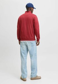 Pullover rosso con colletto alto, vestibilità rilassata, abbinato a jeans azzurri chiari e scarpe beige. Il modello indossa un cappellino blu.
