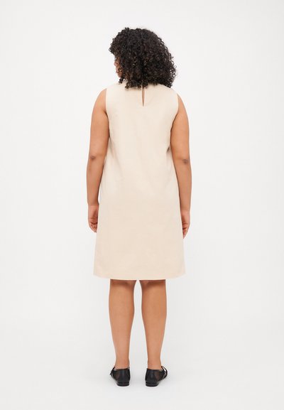 Robe beige sans manches avec un dos en forme de trou de serrure, silhouette droite et texture lisse. Portée avec des chaussures noires.