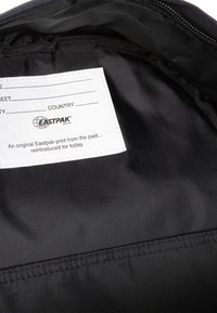 Interior de mochila de tela negra con una etiqueta blanca con campos editables y la marca Eastpak. Textura suave, diseño minimalista.