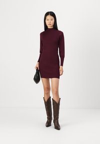 Vínová pletená šaty s dlouhými rukávy, vysokým límcem a slim-fit siluetou. Na sobě má hnědé kovbojské boty a malou černou kabelku.
