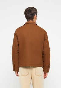 Brown padded jacket with a collar, featuring buttoned cuffs and a straight cut. Worn over light beige pants, showing a simple back view.
