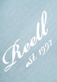 Tela azul clara con texto bordado en blanco que dice "Reell est. 1997" en una fuente cursiva. Se aprecian detalles de la textura de la superficie.