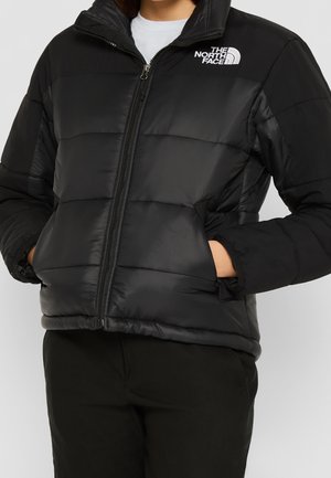 Personne portant une doudoune noire avec un logo blanc "The North Face" sur la poitrine, les mains dans les poches latérales, fermeture éclair fermée.