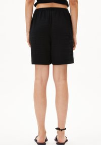 Black shorts made of textured fabric with an elastic waistband, falling to mid-thigh. Worn with flat black sandals and a decorative ankle bracelet.