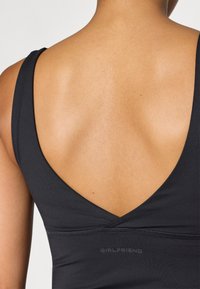Fekete sporttop mély V-hát dizájnnal, sima textúrával, és a "GIRLFRIEND" szó finoman nyomtatva a közepén.