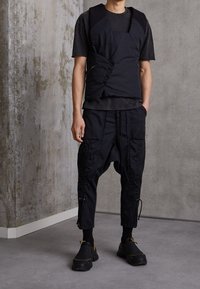 Schwarze, schichtweise Weste über einem dunklen T-Shirt, kombiniert mit weiten schwarzen Hosen mit Cargo-Taschen und Reißverschlüssen, abgerundet mit schwarzen Schuhen.