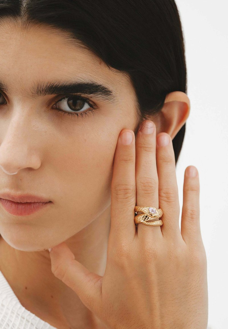 Conjunto de anillos de oro entrelazados con un diseño texturizado y una piedra central, llevados en un dedo contra un fondo neutro.