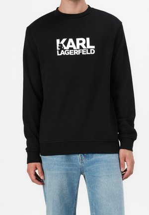 Mann trägt schwarzen Sweatshirt mit langen Ärmeln und weißem "KARL LAGERFELD" Logo sowie hellblaue Jeans, steht vor schlichter Hintergrund.