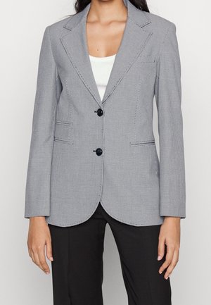 Kvinde iført en tætsiddende grå ternet blazer med sorte knapper over en hvid top, kombineret med sorte bukser.