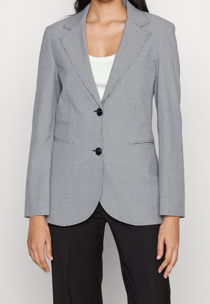 Femme portant un blazer cintré gris à carreaux avec des boutons noirs sur un haut blanc, associé à un pantalon noir.