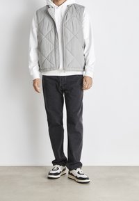 Hellgraue gesteppte Weste über einem weißen Hoodie, kombiniert mit dunklen Jeans und schwarzen sowie cremefarbenen Sneakers. Schlichtes Design mit sichtbaren Steppnähten.