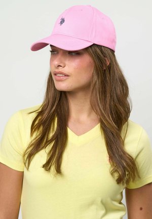 Tânără cu păr lung și castaniu, purtând o șapcă de baseball roz și un tricou galben cu decolteu în V, uitându-se spre stânga.