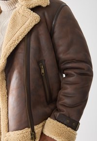 Chaqueta de cuero sintético marrón con un cuello y forro de borrego beige, que cuenta con un bolsillo con cremallera y acentos de puños acolchados.