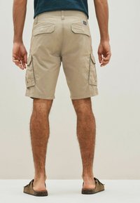 Beige cargo shorts made of cotton blend with a relaxed fit, featuring side and back pockets, and rolled hems. Worn with brown slide sandals.