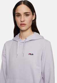 Huppari vaalean lilassa värissä, joka on valmistettu pehmeästä kankaasta. Siinä on etupuolella kenguru-tasku, narut hupussa ja punaisella sekä sinisellä brodeerattu Fila-logo.