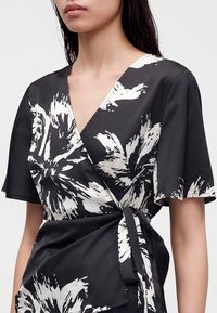 Jeune femme portant une robe portefeuille fleurie noire et blanche à manches courtes avec une ceinture nouée à la taille.