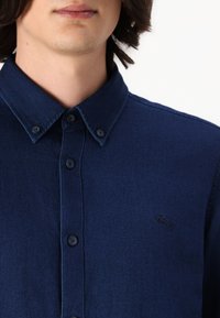 Primo piano di una persona che indossa una camicia blu scuro abbottonata con colletto e piccolo logo ricamato sul petto.