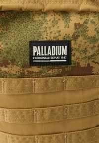 Batoh Palladium s maskovacím vzorom v zelených a hnedých farbách, s čiernou logo nálepkou a béžovými nylónovými popruhmi a akcentmi.