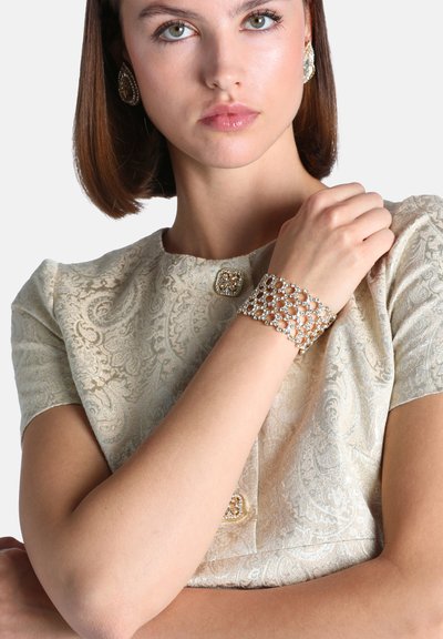 Jeune femme aux cheveux bruns lisses porte une robe crème à motifs et un bracelet orné en or et strass ainsi que des boucles d'oreilles assorties.