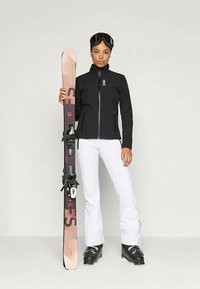 Schwarze Skijacke mit Reißverschluss, weiße Skihose, schwarze Skischuhe und pinke Skier mit schwarzen Bindungen sowie eine Skibrille auf dem Kopf.