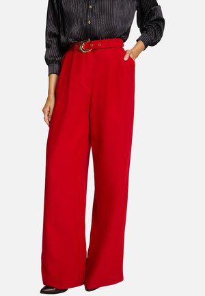 Pantalons larges rouges en tissu lisse, avec une ceinture à boucle dorée et des poches latérales. Associés à un chemisier à rayures noires.