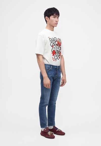 Jeune homme debout, portant un t-shirt graphique blanc, un jean slim bleu, des chaussettes blanches et des baskets rouges sur un fond blanc uni.
