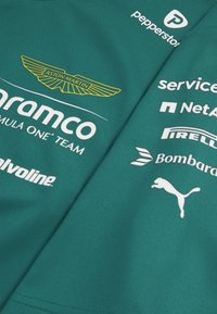 Teal kortærmet trøje med Aston Martin-logoet i guld. Inkluderer hvide sponsorlogoer: Aramco, Pirelli, Bombardier og Puma.