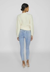 Jersey de punto crema con patrón texturizado, combinado con unos jeans ajustados de un azul claro. La modelo lleva tacones beige, con el cabello recogido en un moño bajo.