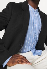 Blazer di lana nera con rever a punta, indossato sopra una camicia a righe azzurre e bianche con bottoni, abbinato a pantaloni crema. Dettagli del tessuto texturizzati evidenti.