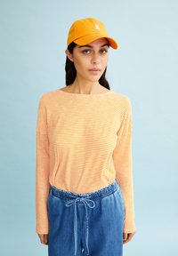 Gestreepte lange mouwen top in oranje en wit, gecombineerd met blauwe broek met trekkoord. De pet is oranje met een wit logo. Zachte stoftextuur.