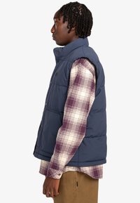 Navy blue puffer vest with high collar, front buttons, and side pockets, layered over a plaid shirt in white and purple tones.