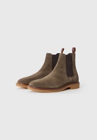 Par de botas Chelsea de ante marrón con paneles laterales elásticos oscuros y suelas de goma color goma sobre un fondo blanco.