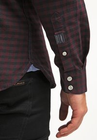 Camicia a maniche lunghe a fantasia quadri bordeaux e nera con polsini abbottonati, dotata di un'etichetta vicino al braccio e di un accento in tessuto blu all'orlo.