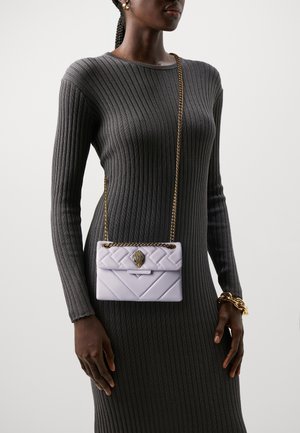 Lyslilla quiltet håndtaske med guld kædetræk, der har en dekorativ lås, holdt af en person iført en grå ribbet langærmet kjole.