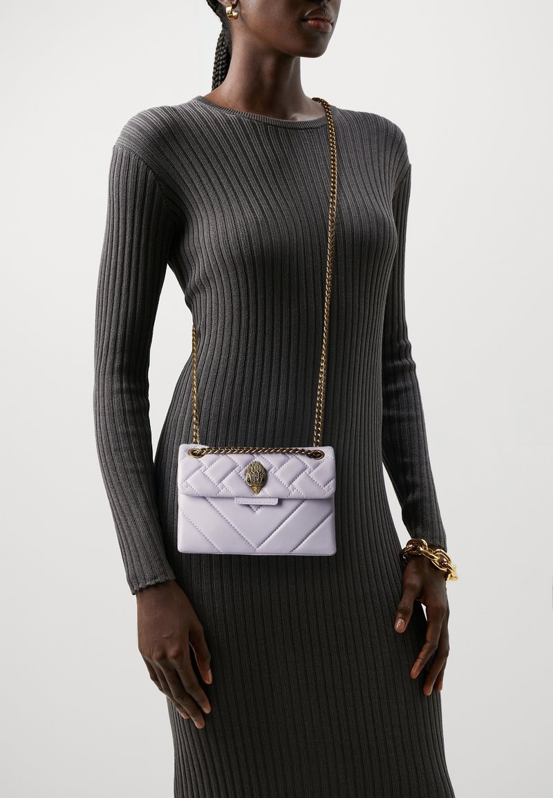 Svetle fialová prošívaná kabelka se zlatým řetízkovým popruhem, s dekorativními sponami, kterou drží osoba oblečená v šedých žebrovaných dlouhých šatech.