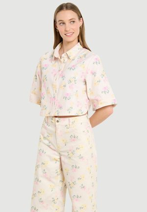 Młoda kobieta ubrana w kremową, kwiatową, krótką koszulę na guziki oraz szerokie, wysokodupiaste spodnie, uśmiecha się i patrzy w prawo.