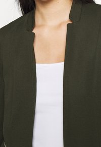 Olivgrön ribbad jacka med hög krage och öppen framdesign, lager på lager över en vit tank topp. Slät textur med minimal detaljer.