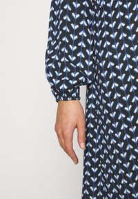 Manche et main d'une personne portant un tissu noir avec un motif géométrique bleu et blanc, avec un poignet élastique froncé.