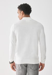 Suéter blanco de punto con dobladillo y puños acanalados, que presenta un diseño simple. Combinado con jeans grises, mostrando un ajuste casual.