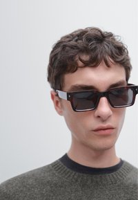 Occhiali da sole con forma rettangolare, lenti scure, montatura tartarugata e dettagli bianchi. Indossati da una persona con capelli corti e ricci.