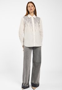 Langärmliges weißes Hemd mit schwarzen Perlenverzierungen, die über die Vorderseite herabfallen, kombiniert mit grauen Jeanshosen im weit geschnittenen Stil und beigen Absätzen.