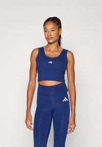 Tamsiai mėlynas trumpas viršus, pagamintas iš minkštos medžiagos, su apvaliu iškirpimu ir baltu Adidas logotipu, derinamas su aukštu liemeniu tamsiai mėlynomis tamprėmis, turi baltas juosteles.