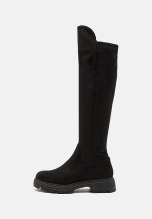 Bottes montantes en daim noir avec une semelle en caoutchouc plate, un bout arrondi et un détail de couture. Le haut présente un léger revers.