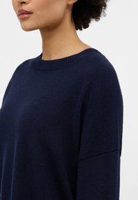 Dunkelblaues Pulli mit geripptem Rundhalsausschnitt, weicher Textur und lockerer Passform; sichtbare Nähte entlang der Nähte.